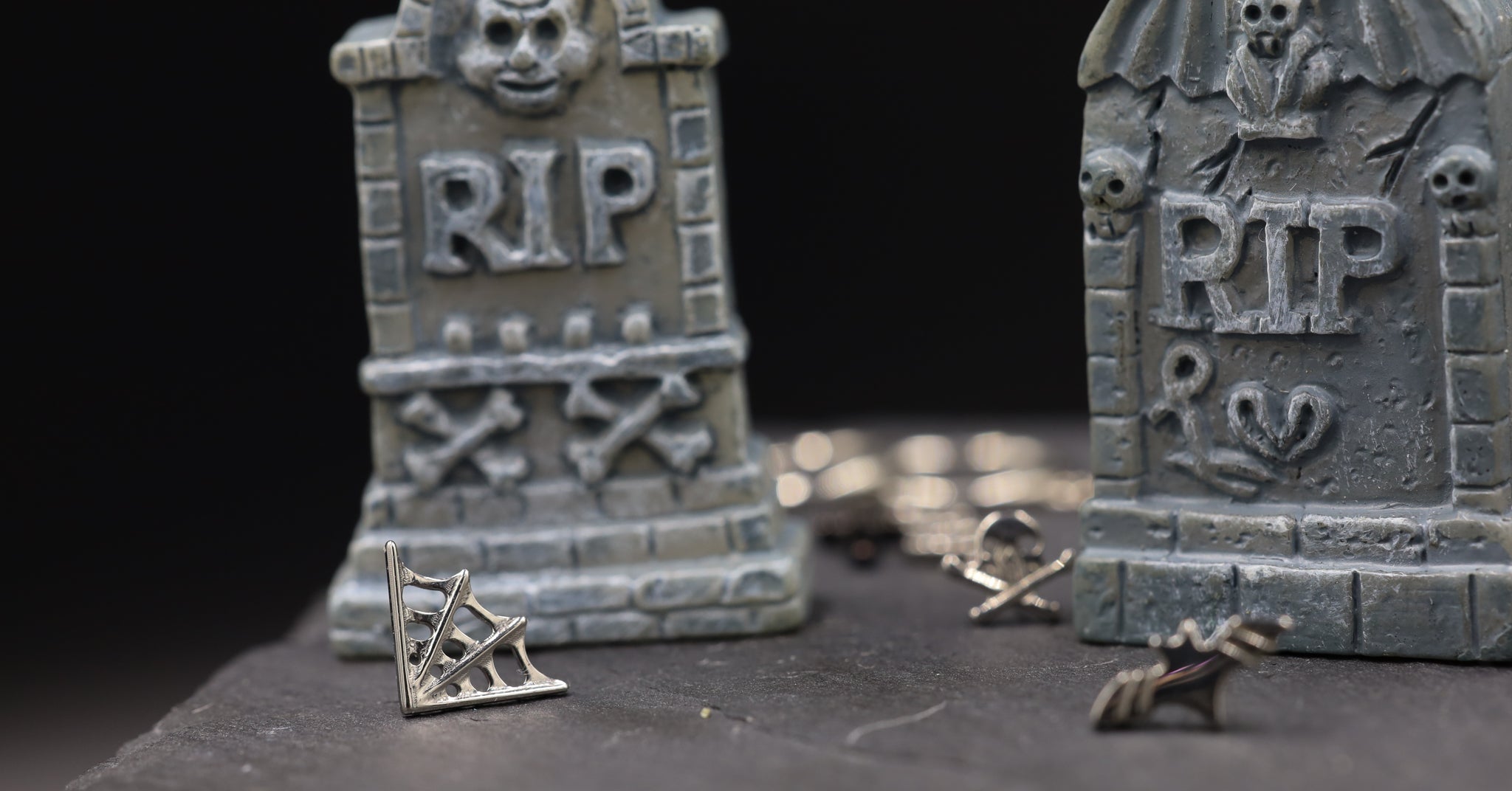 Two miniature tombstones with 'RIP' on a dark background with titanium cobweb and bat earring in the foreground